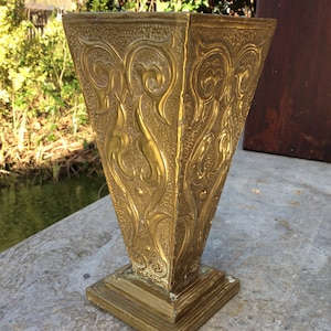 May include: A gold metal vase with an intricate, embossed floral design. The vase has a square base and a wide, flared opening.