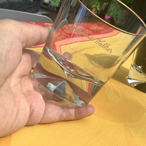 May include: Clear, faceted whiskey glass with the text "Johnnie Walker" etched on the side. The glass is held in a hand and is partially filled with a clear liquid. A second, dark glass is in the background.