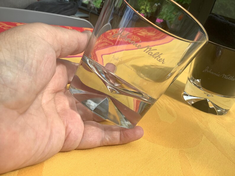 May include: Clear, faceted whiskey glass with the text "Johnnie Walker" etched on the side. The glass is held in a hand and is partially filled with a clear liquid. A second, dark glass is in the background.