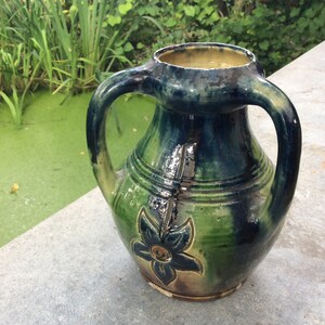 Può includere: Un vaso in ceramica verde e blu con due manici e un motivo floreale. Il vaso è appoggiato su una superficie di cemento.