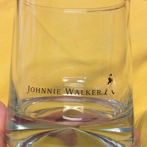 May include: A clear glass tumbler with the words "JOHNNIE WALKER" printed on the side. A small silhouette of a man walking with a cane is printed on the bottom right side of the glass.