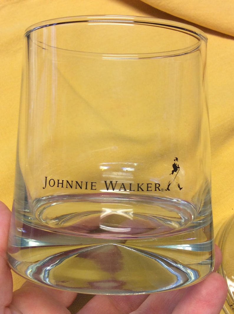May include: A clear glass tumbler with the words "JOHNNIE WALKER" printed on the side. A small silhouette of a man walking with a cane is printed on the bottom right side of the glass.
