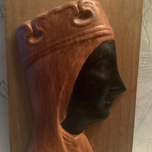 Puede incluir: Una placa de pared de madera con un perfil tallado de una persona que lleva una corona y una túnica con capucha. La cara de la persona es negra y la túnica es marrón.