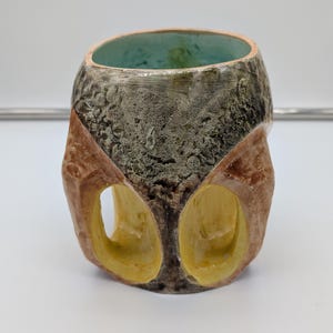 May include: A ceramic planter with a textured, gray, brown, and yellow glaze. The planter has two open, oval-shaped cutouts on the side.