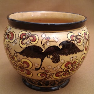 Può includere: Un vaso in ceramica con un bordo nero e un corpo color crema. Il vaso è decorato con un disegno di un uccello nero e motivi floreali rossi, gialli e bianchi.