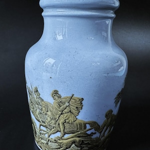 Könnte beinhalten: Eine hellblaue Keramikvase mit einem goldenen Design einer Jagdszene. Die Szene zeigt einen Mann zu Pferd mit einem Hund, der neben ihm läuft.