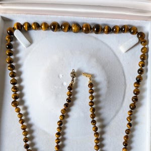 Peut inclure: Un collier en or avec un fermoir en perles de œil de tigre. Le collier est présenté dans une boîte blanche.