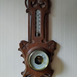 May include: A wooden wall-mounted barometer and thermometer with a round face and a carved frame. The barometer face has the text "MPESTA PONGSIA VARIABILE BELIMO SENSECO". The thermometer has a scale in degrees Celsius.