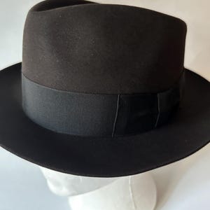Op de afbeelding: Een zwarte fedora hoed met een grosgrain lint. De hoed heeft een klassiek ontwerp met een brede rand en een gestructureerde kroon. De hoed wordt getoond op een witte mannequin hoofd.