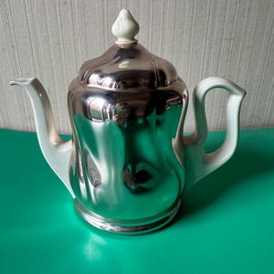 Op de afbeelding: Een zilveren theepot met een wit deksel en handvat. De theepot heeft een glanzend, reflecterend oppervlak en een rond lichaam.