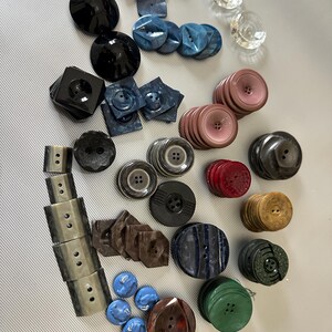 Può includere: Una collezione di bottoni vintage in vari colori, forme e dimensioni. I bottoni sono realizzati in plastica, metallo e legno. Alcuni dei bottoni hanno un design decorativo, mentre altri sono semplici. I bottoni sono disposti in uno schema casuale su una superficie bianca.
