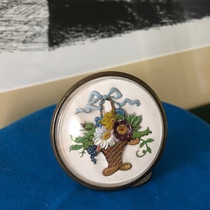 Puede incluir: Un pequeño adorno redondo de vidrio con fondo blanco y un diseño floral colorido. El diseño presenta una cesta llena de flores y una cinta azul.