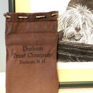 Può includere: Una borsa di tela marrone con chiusura a coulisse e il testo "Durham Trust Company Durham, N.H." stampato sul davanti. La borsa è accanto a un disegno in bianco e nero incorniciato di un cane in un passeggino.