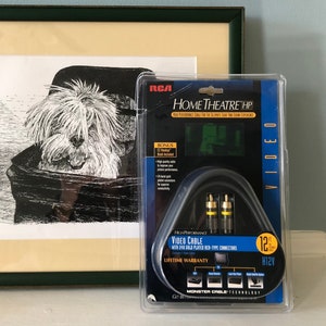 May include: A black and white line drawing of a dog in a basket, framed in a green frame. Next to the frame is a package of RCA Home Theatre HP 12' video cable with 24K gold-plated RCA-type connectors. The package is blue and black with the RCA logo and the text "Home Theatre HP" and "Video Cable".