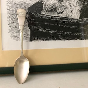 以下が含まれることがあります： 装飾的な柄の付いた銀のスプーンが、犬の白黒の額縁入り絵画の前にあるベージュの表面に置かれています。