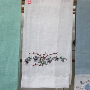 Op de afbeelding: Witte linnen theedoek met geborduurde paarse bloemen en groene bladeren. De handdoek hangt aan een houten rek met twee andere handdoeken, een lichtblauwe en een lichtgroene.