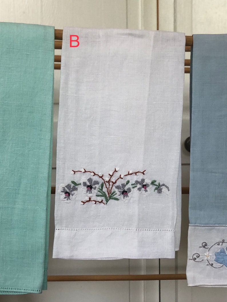 Op de afbeelding: Witte linnen theedoek met geborduurde paarse bloemen en groene bladeren. De handdoek hangt aan een houten rek met twee andere handdoeken, een lichtblauwe en een lichtgroene.
