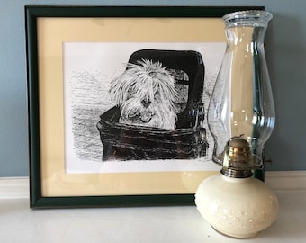 May include: A black and white pen and ink drawing of a dog in a basket, framed in a dark green frame. A white oil lamp with a clear glass chimney sits on a white surface next to the frame.