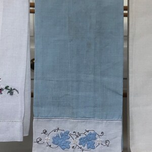 Op de afbeelding: Een lichtblauwe theedoek met een witte rand en geborduurde blauwe bladeren. De handdoek hangt aan een houten rek.