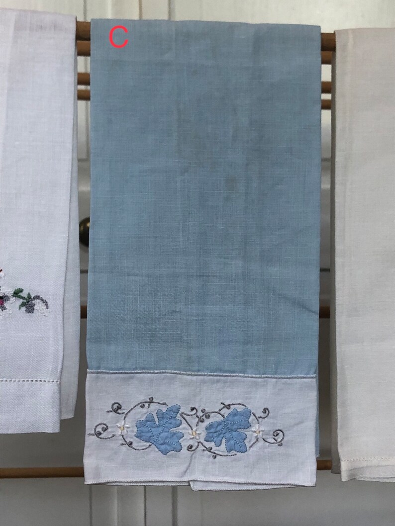 Op de afbeelding: Een lichtblauwe theedoek met een witte rand en geborduurde blauwe bladeren. De handdoek hangt aan een houten rek.