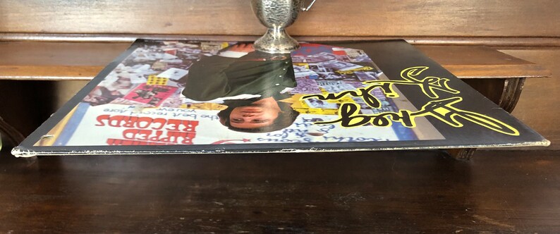 Op de afbeelding: Een zwart vinyl-muziekalbum met een kleurrijke collage van afbeeldingen op de hoes. De hoes van het album toont een man die een groene jas en een geel shirt draagt. De titel van het album is "The Rod Stewart Album" in gele letters.