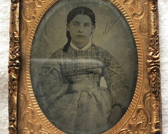 May include: A black and white portrait of a young woman in a plaid dress. The photo is framed in a gold oval frame with ornate details.