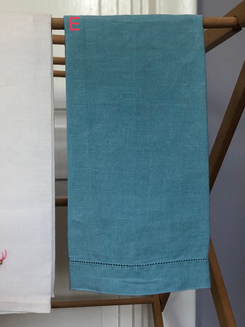 Op de afbeelding: Een lichtblauwe linnen theedoek met een witte geborduurde rand. De handdoek hangt aan een houten droogrek.