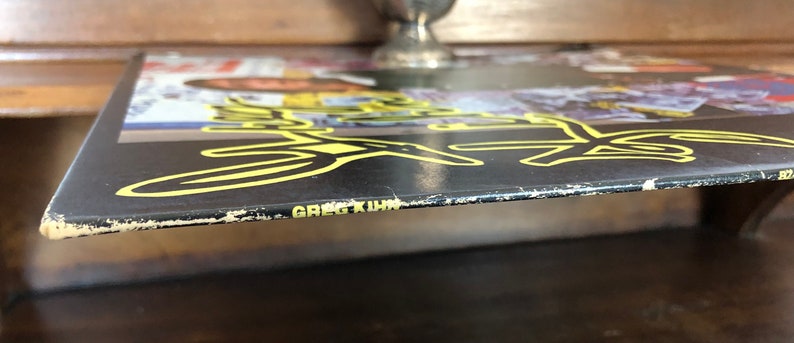 Op de afbeelding: Een zwarte vinylplaat met een geel en zwart ontwerp. De plaat is getiteld "Greg Kihn".