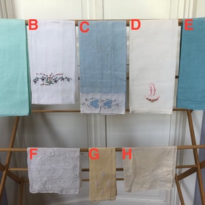Op de afbeelding: Acht geborduurde handdoeken hangen aan een houten droogrek. De handdoeken zijn in verschillende kleuren, waaronder wit, lichtblauw en beige. Sommige handdoeken hebben bloemenborduurwerk, terwijl andere een enkele letter hebben.