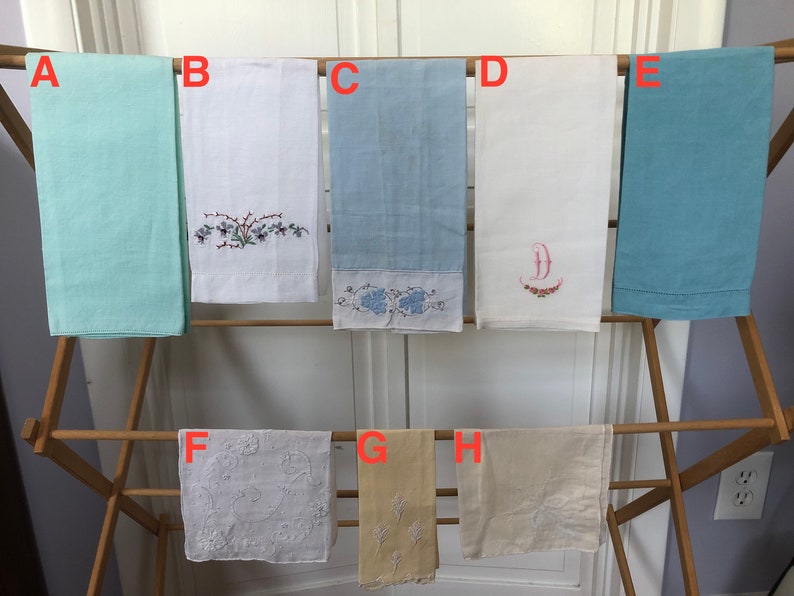 Op de afbeelding: Acht geborduurde handdoeken hangen aan een houten droogrek. De handdoeken zijn in verschillende kleuren, waaronder wit, lichtblauw en beige. Sommige handdoeken hebben bloemenborduurwerk, terwijl andere een enkele letter hebben.