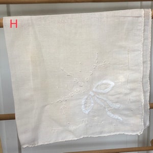 Op de afbeelding: Een wit linnen zakdoek met een delicaat geborduurd bloemenmotief. De borduurwerk toont een witte strik met een bloeiende stengel en bladeren.