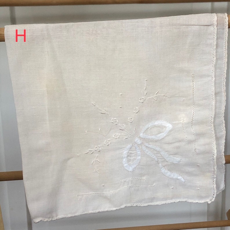Op de afbeelding: Een wit linnen zakdoek met een delicaat geborduurd bloemenmotief. De borduurwerk toont een witte strik met een bloeiende stengel en bladeren.