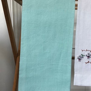Op de afbeelding: Een lichtblauwe linnen theedoek die aan een houten droogrek hangt. De handdoek heeft een delicate kantversiering rond de randen.