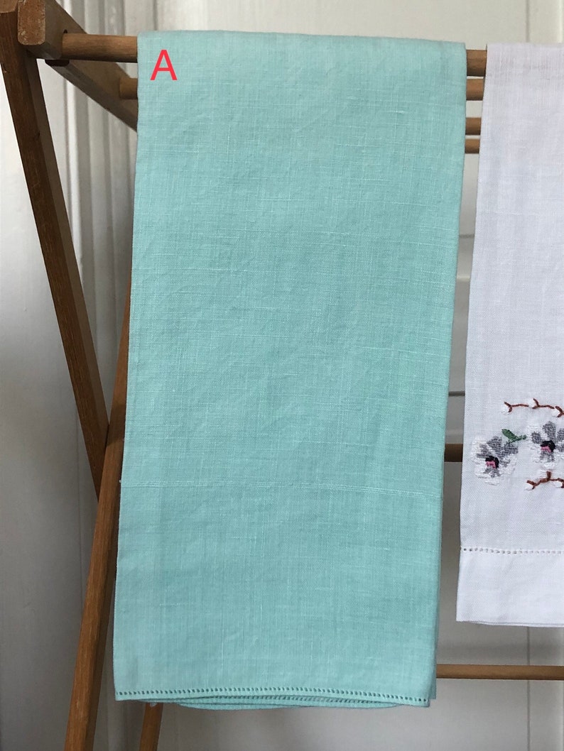 Op de afbeelding: Een lichtblauwe linnen theedoek die aan een houten droogrek hangt. De handdoek heeft een delicate kantversiering rond de randen.