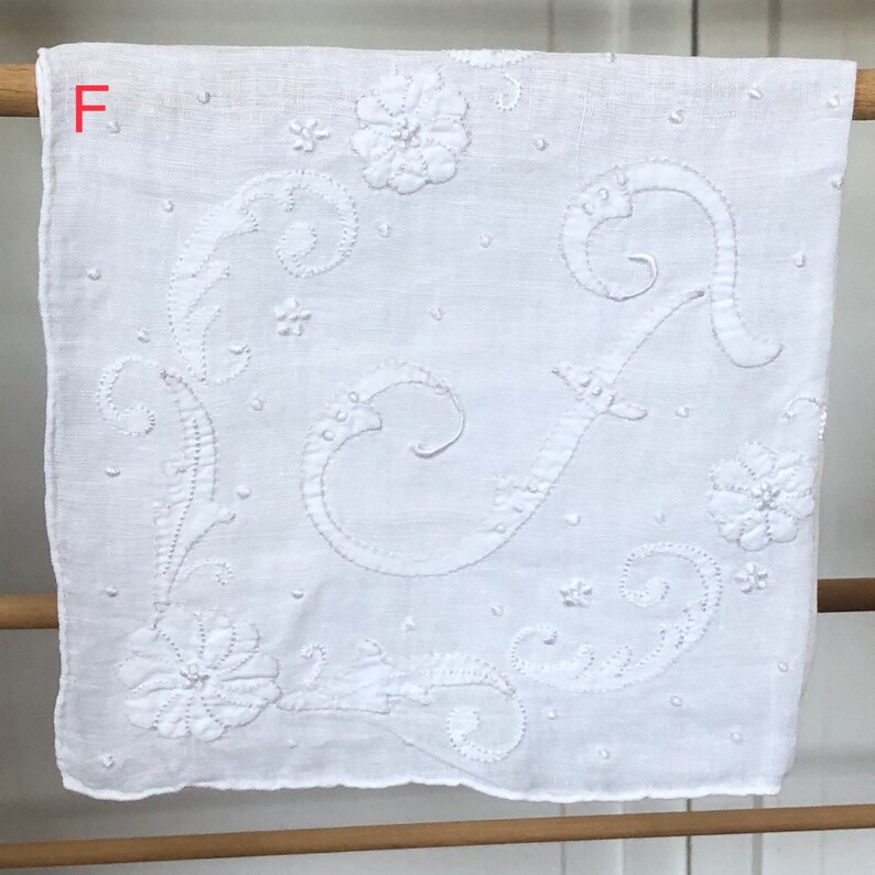 Op de afbeelding: Wit katoenen zakdoek met delicate witte borduursels van een bloemenmotief en de letter "F".
