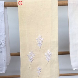 Op de afbeelding: Een gele linnen theedoek met witte geborduurde bloemenmotieven. De handdoek heeft een gegolfde rand en de letter "G" is zichtbaar in de bovenhoek.