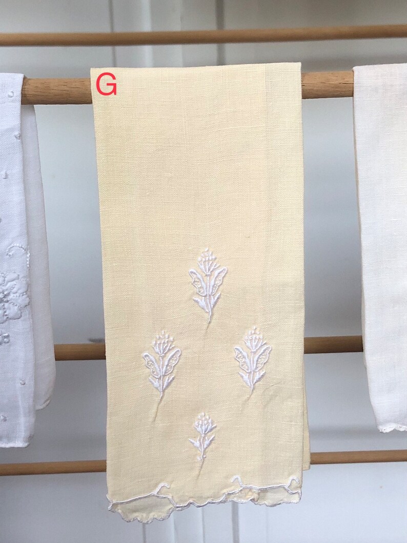 Op de afbeelding: Een gele linnen theedoek met witte geborduurde bloemenmotieven. De handdoek heeft een gegolfde rand en de letter "G" is zichtbaar in de bovenhoek.
