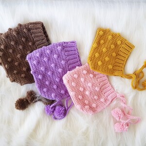 May include: Four crocheted baby hats with pom-poms. The hats are in brown, purple, pink, and yellow.