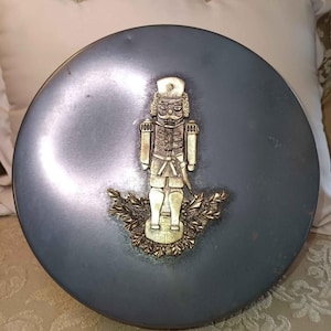May include: A round, blue-grey decorative box with a gold-colored nutcracker figure on the lid. The nutcracker is standing on a gold-colored wreath. The box is sitting on a patterned surface.