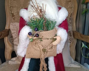 May include: A Santa Claus figurine with a white beard, blue eyes, and a red velvet coat trimmed with white faux fur. He holds a burlap sack filled with greenery and berries, adding a festive touch to the holiday decor.