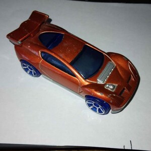 May include: A bright orange toy car with a blue windshield and blue wheels. The car has a large rear wing and a detailed engine.