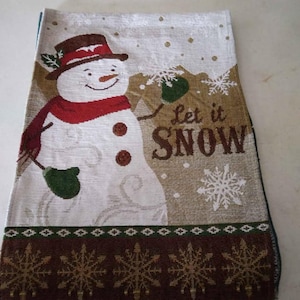 May include: A rectangular Christmas table runner featuring a smiling snowman wearing a red scarf and a brown hat. The words "Let it Snow" are printed on the runner, along with snowflakes. The bottom border has a repeating snowflake pattern.