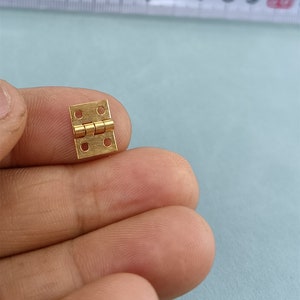 May include: A small gold-colored metal hinge with four holes on each side. The hinge is closed and appears to be made of brass.