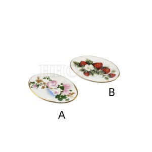 Może przedstawiać: Dwa owalne ceramiczne talerze z pozłacanym brzegiem. Jeden talerz przedstawia wzór kwiatowy z różowymi różami i zielonymi liśćmi. Drugi talerz przedstawia wzór z czerwonymi truskawkami i zielonymi liśćmi. Talerze są oznaczone literami A i B.