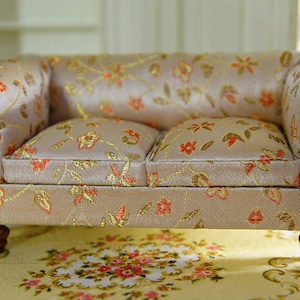 May include: A miniature two-seater sofa with a beige floral pattern. The sofa has a wooden frame with dark brown accents and turned legs. The cushions and back are covered in a fabric with gold, orange, and green floral designs.