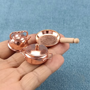 Puede incluir: Un juego de utensilios de cocina de cobre en miniatura, que incluye una tetera, una olla con tapa y una sartén con mango de madera.