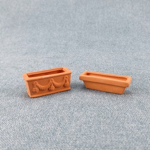 Puede incluir: Dos macetas de terracota, una con un diseño decorativo y la otra lisa, sobre una superficie azul texturizada.