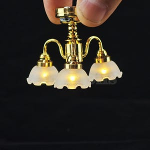 May include: Miniature gold chandelier with three frosted glass shades. The small light fixtures are illuminated, creating a warm glow. The chandelier is held by a hand against a black background.