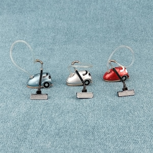 以下が含まれることがあります： 黒いホースとハンドルが付いた、赤、銀、青のミニチュアのおもちゃ掃除機が3つ。掃除機は青い表面の上に置かれています。