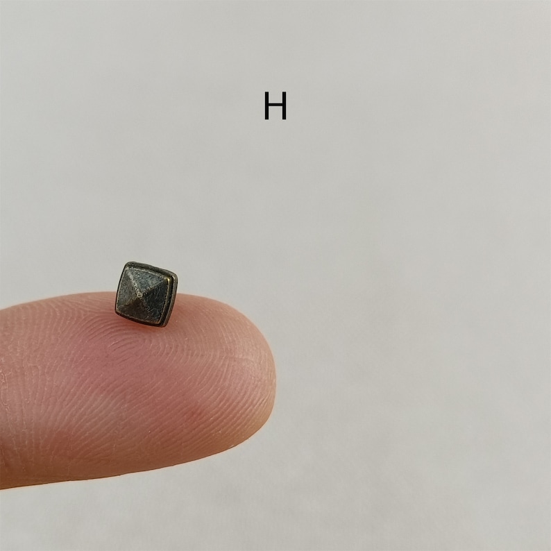 Puede incluir: Un peque&ntilde;o remache de metal cuadrado de forma piramidal con un acabado gris oscuro. El remache se sostiene en un dedo.
